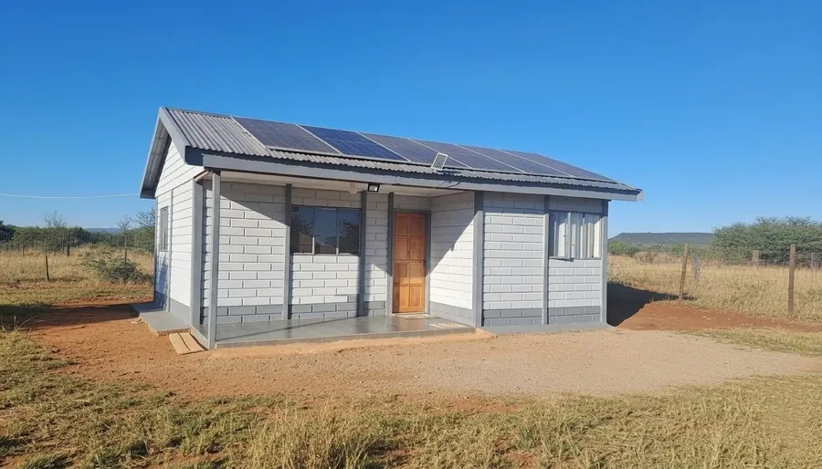 Precast two-bedroom house with a covered stoep and solar panels on the roof, standing on open veld